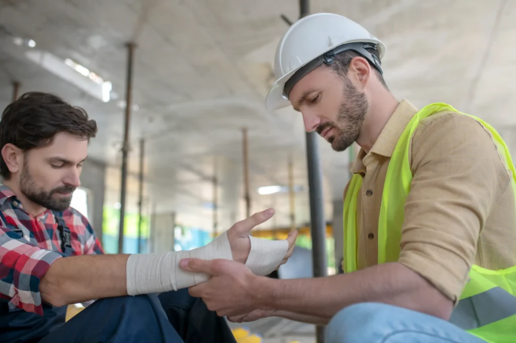 Construction worker in a helmet applying bandage on his coworker's arm.